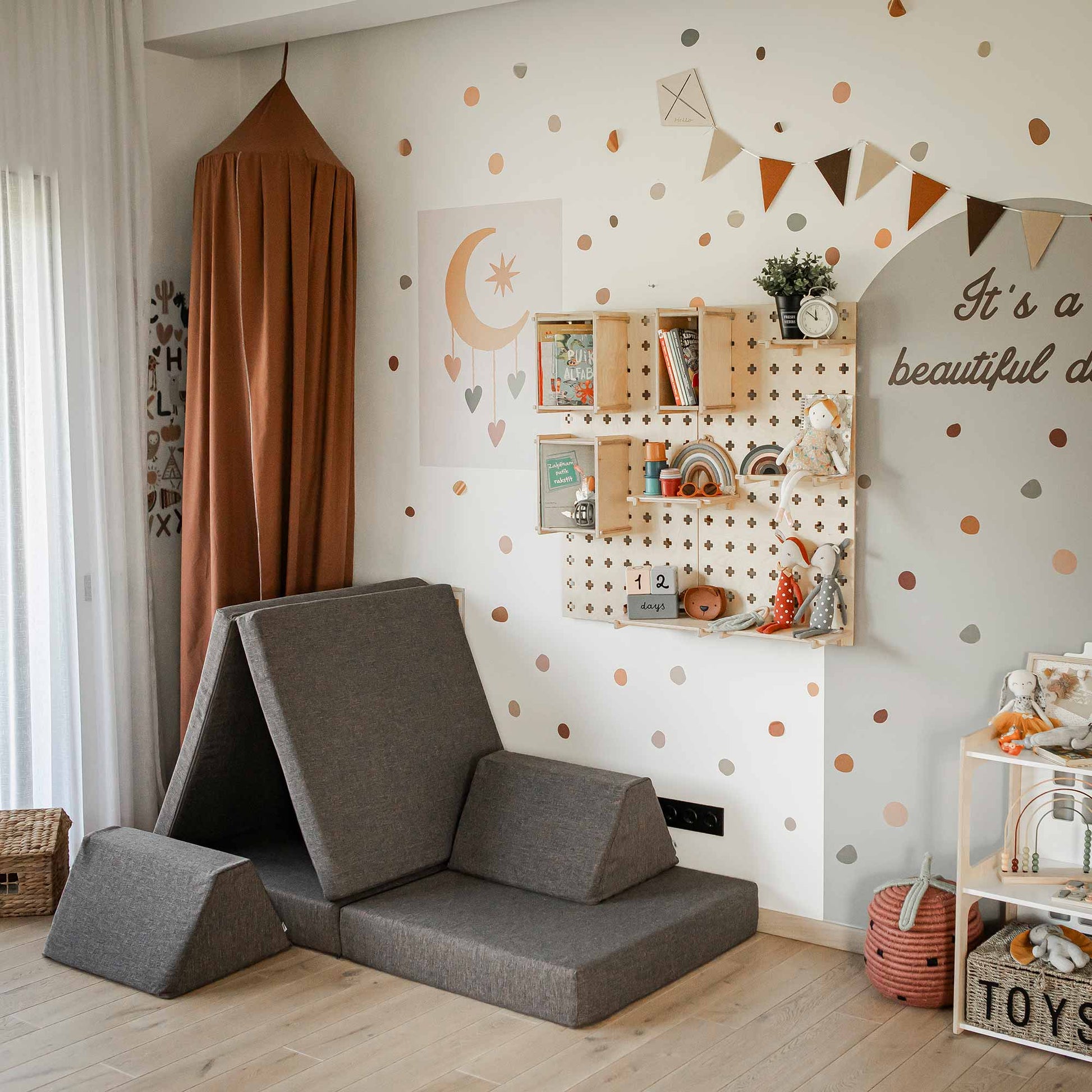 A children's playroom with colorful wall decor, a hanging canopy, shelves with toys, and a Monboxy Mattress set - Dark brown featuring foldable gray cushions. A banner reads "It's a beautiful day," inviting open-ended play and fostering gross motor skills.