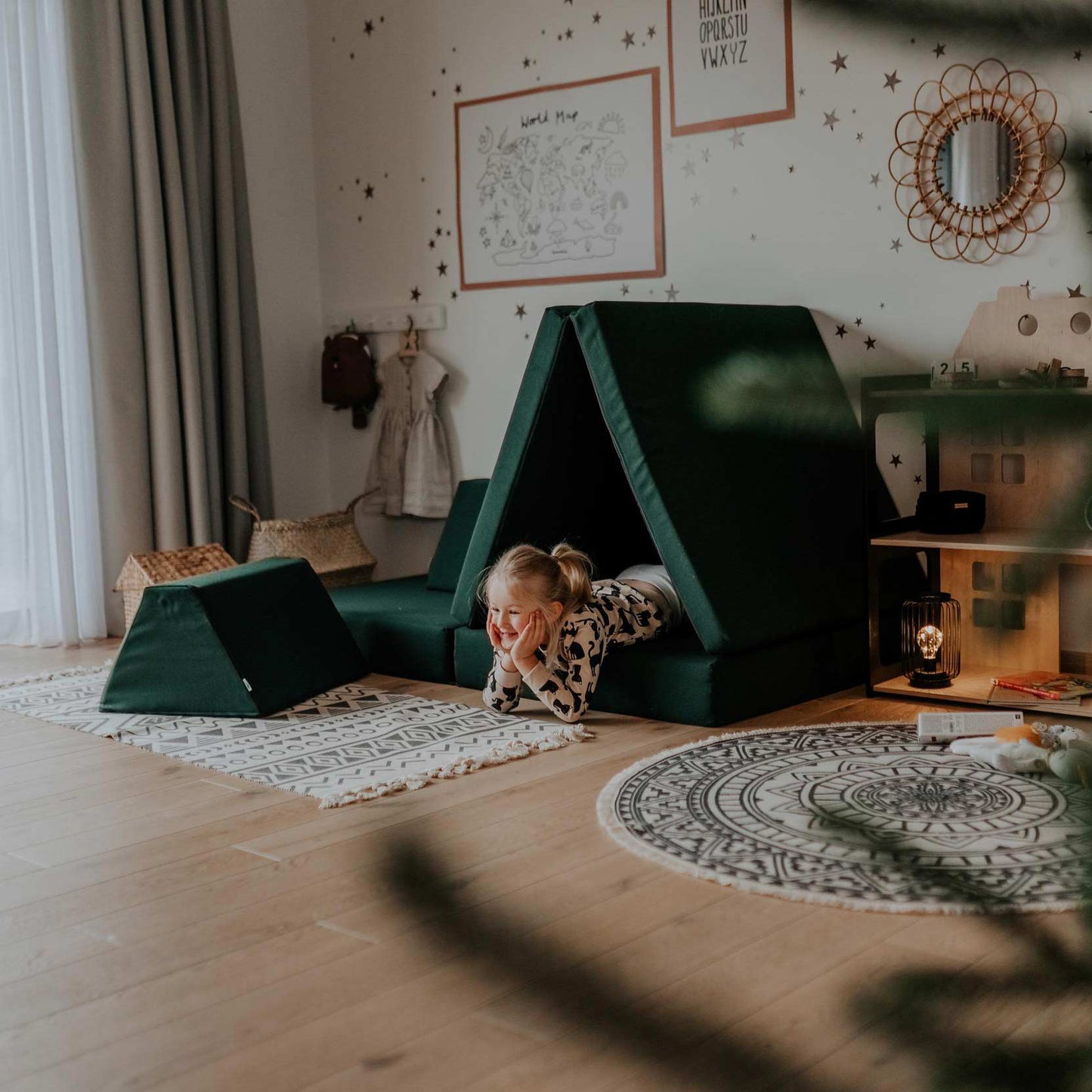 A young girl plays on a Monboxy Play Mattress in Deep Green inside a cozy Montessori room decorated with children's artwork, soft lighting, and a patterned rug.