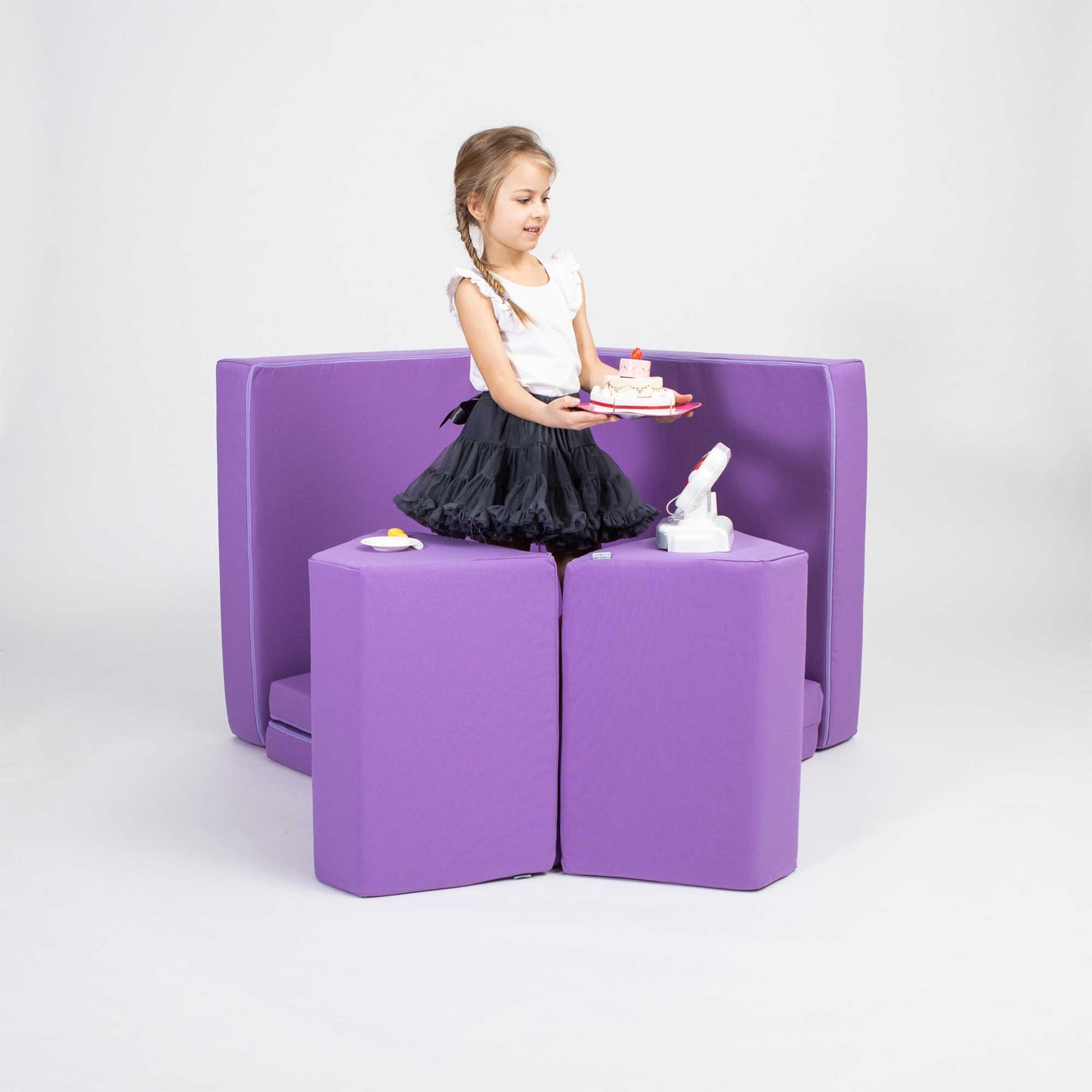 A young girl in a black dress plays with a Monboxy soft play mattress set in purple on a Monboxy foam play couch in a studio setting.