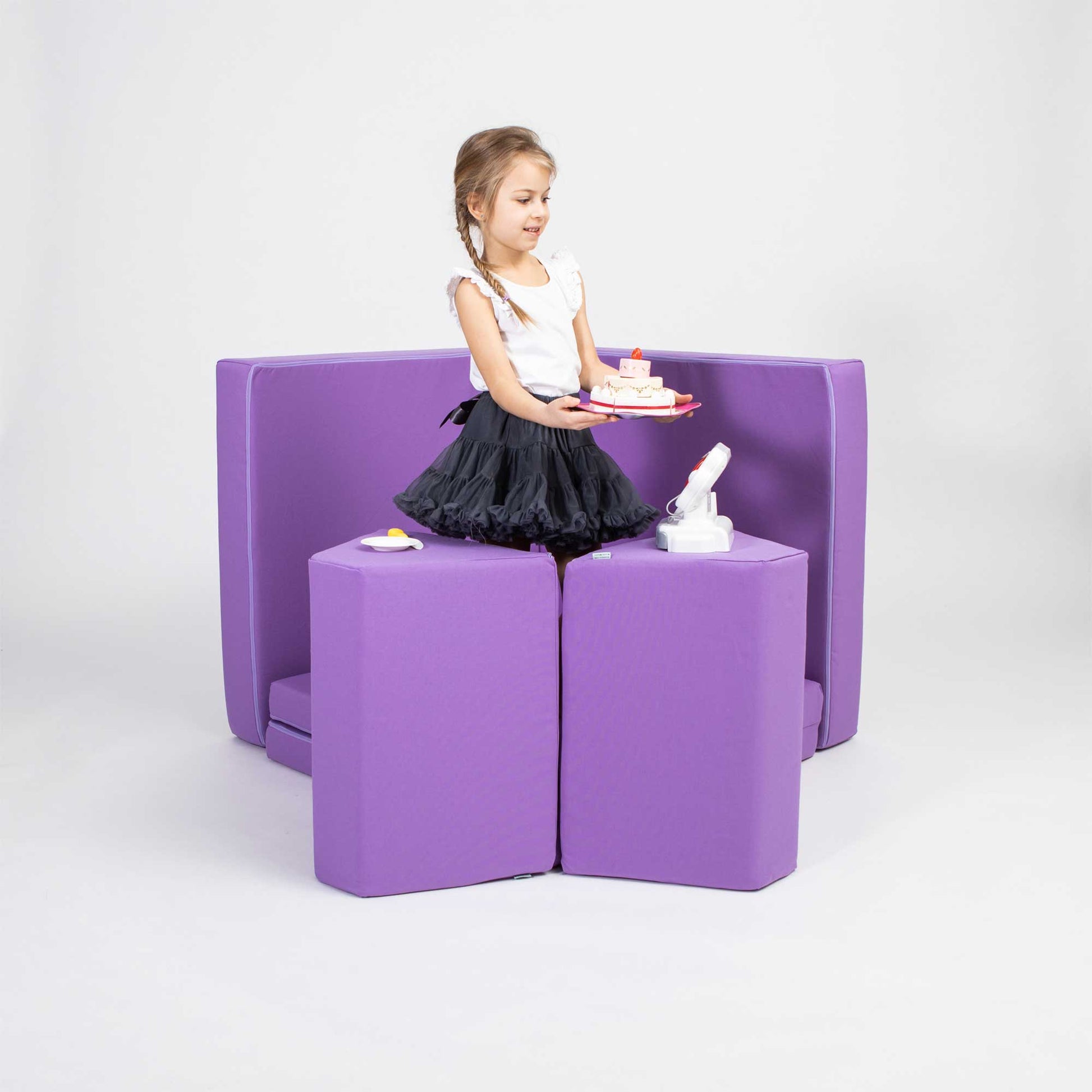 A young girl in a black dress plays with a Monboxy soft play mattress set in purple on a Monboxy foam play couch in a studio setting.