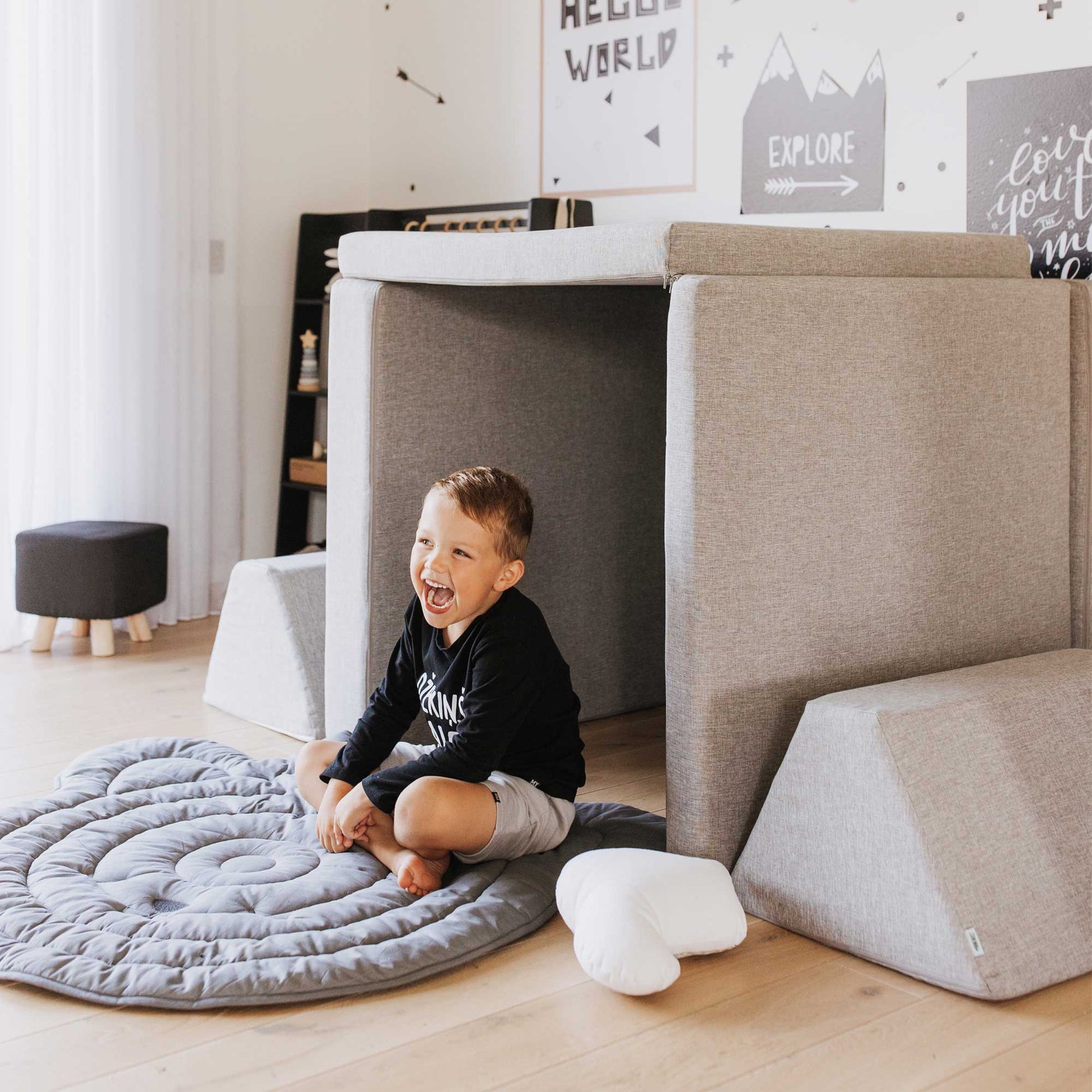 A toddler boy laughing sitting on a grey rug next to his Monboxy play couch