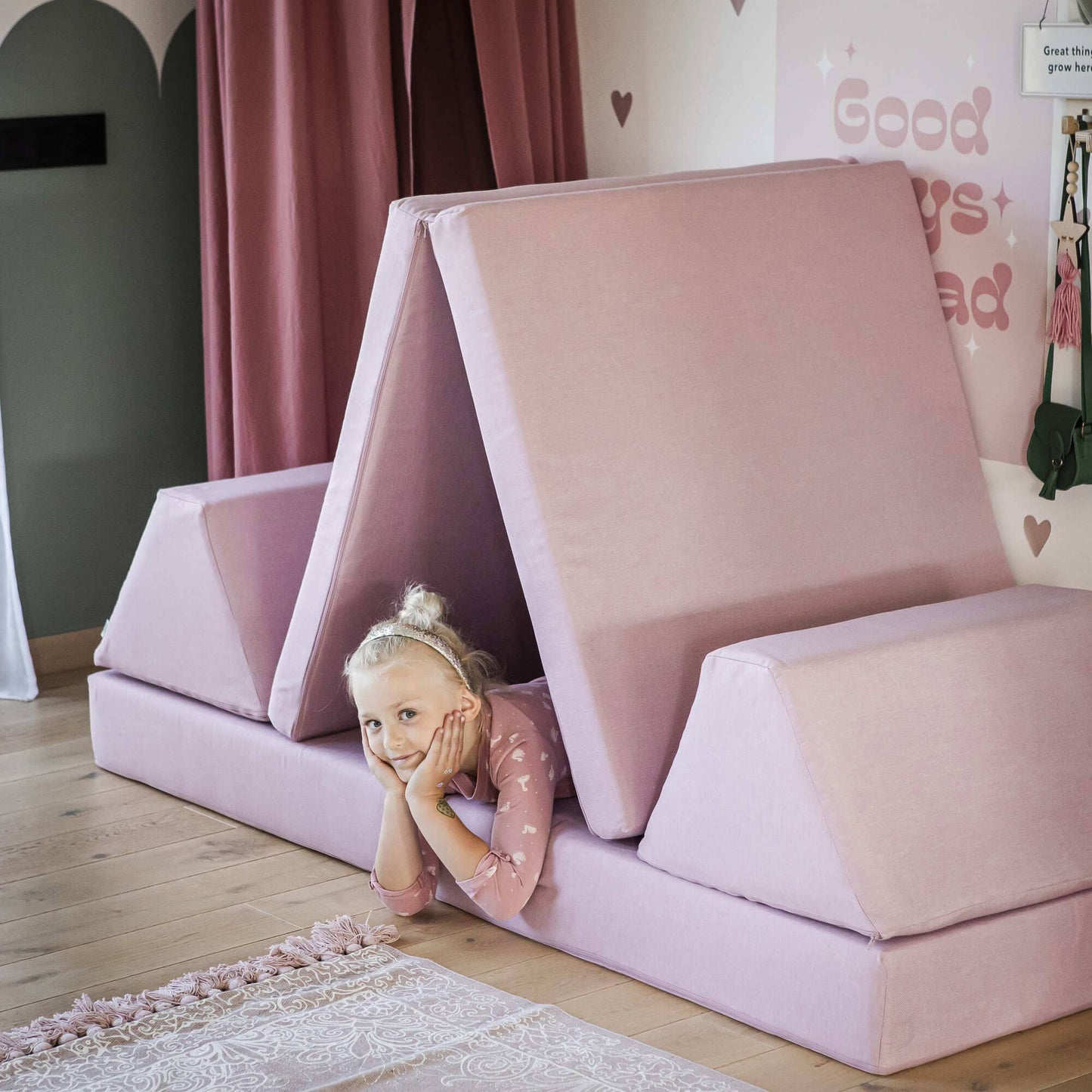 A child beams with joy under a Monboxy Foam play mattress set - Pink, cleverly shaped like a tent on wooden flooring. This cozy setup doubles as an indoor play gym, making it the perfect Montessori gift.