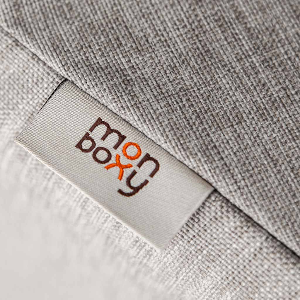 Close-up of a gray fabric texture with a white label stitched on it, displaying the logo "Monboxy" in lowercase letters with an orange cross above the letter 'n'.