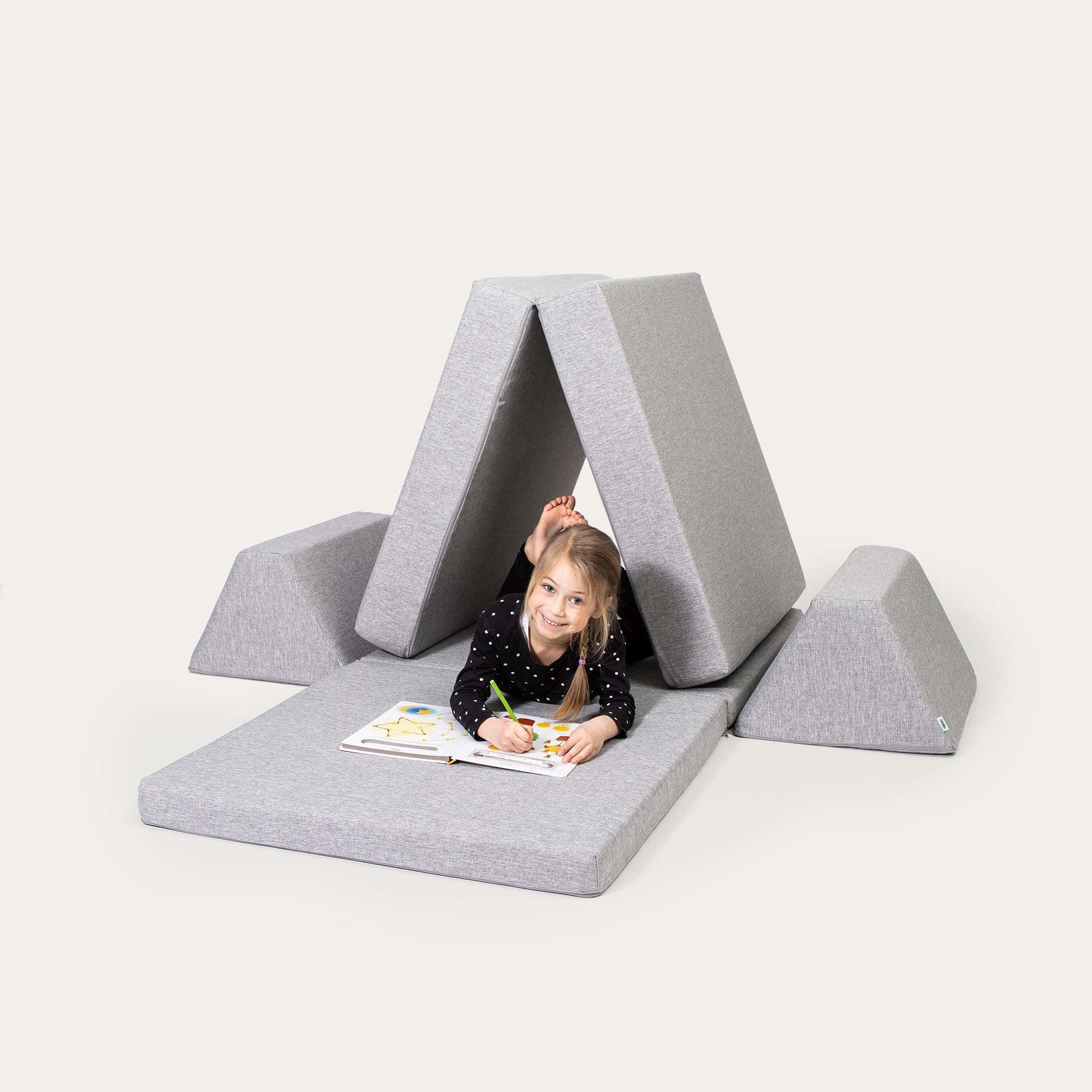 A little girl drawing in a coloring book while sleeping on a grey triangle-shaped Monboxy activity sofa