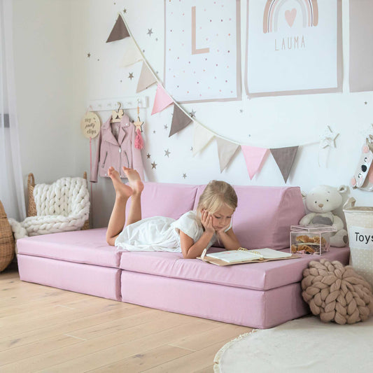 A girl laying on her tummy reading a book on her pink Monboxy play sofa