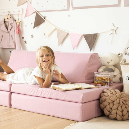 Young girl lying on a Monboxy pink foam play mattress set in a playroom, reading a book and smiling, surrounded by toys and decorative items.