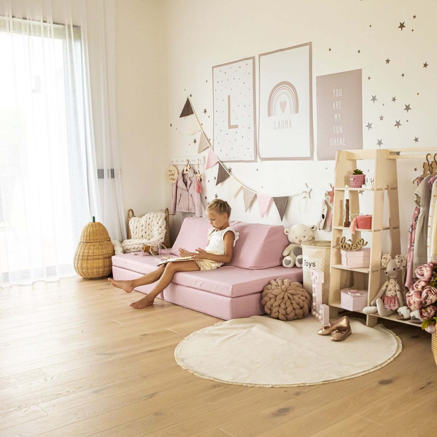 Young girl sitting on a Monboxy pink foam play mattress set in a playroom, reading a book and smiling, surrounded by toys and decorative items.